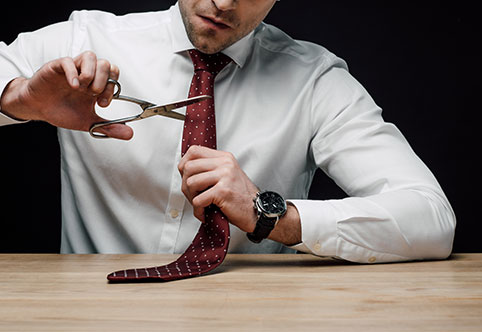image of man sitting at a desk wearing a white button up shirt and holding a pair of scissors and cutting the dark red tie around is neck
