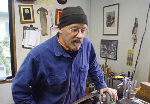 image of a man wearing a dark blue button up flannel shirt and a brown knit cap working with metal in a workshop